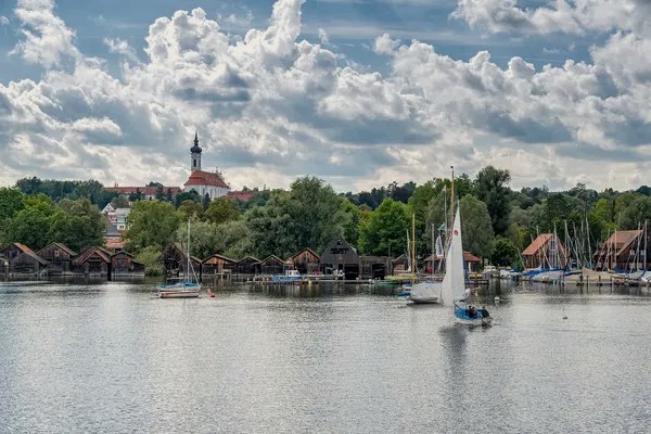 A sailing boat on Lake Starnberg in front of boathouses and a church on the shore